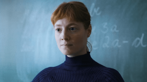 a young woman with red hair, wearing a black polo neck jumper, stands in front of a classroom chalk board 