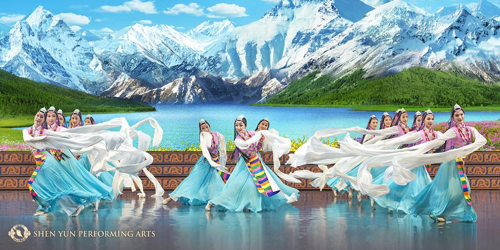 Image of traditional Chinese dancers, wearing blue and white outfits. The background features a lake and snow-topped mountains. 