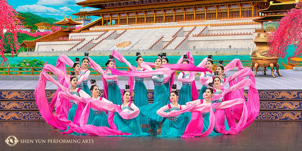Image of traditional Chinese dancers, wearing blue and pink outfits. The background features a Chinese temple. 