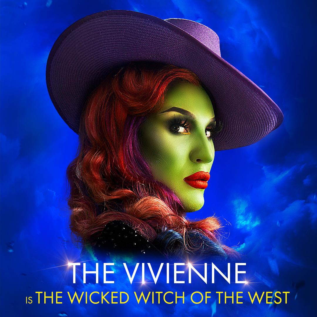The Vivienne with a green face, long red hair and wearing a purple, wide-brimmed hat. Against a dark blue, cloudy backdrop.