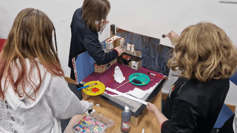 three children with their backs to the camera set up a stop-motion animation set, on a Halloween theme 
