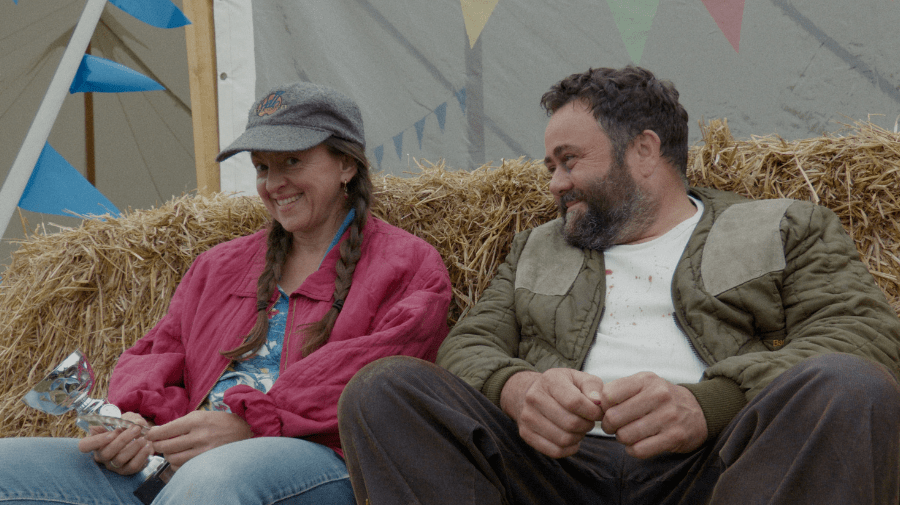 two people sit against hay bales. the woman has long hair in braids and wears a baseball cap,she's holding a small silver awards cup.The man is in a dirty t-shirt and trousers and smiles at the woman 
