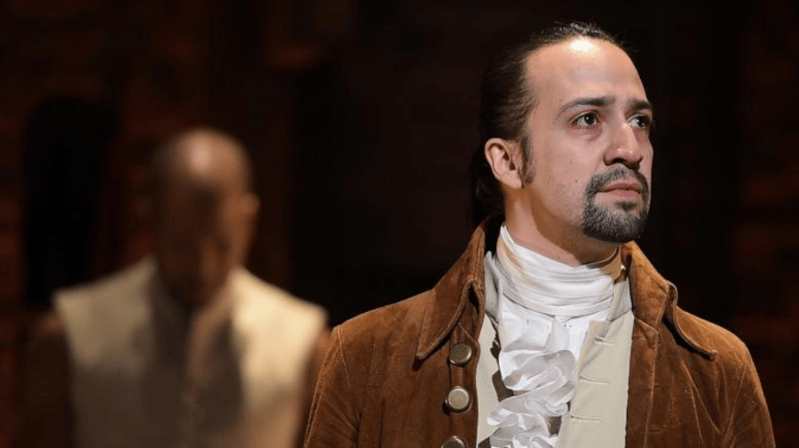 a man with slicked back,black hair, in a ponytail and a dark goatee beard, is dressed in a brown coloured frock coat and cream ruffled shirt, as worn in the 18th Century. He's looking off into the distance. Behind him, the blurred image of another man in a cream frock coat
