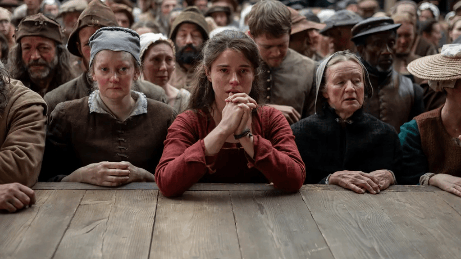 a woman in a 16th century deep red dress, stands wit her elbows on the edge of a stage. she is surrounded by the rest of the audience, all in similar dress
