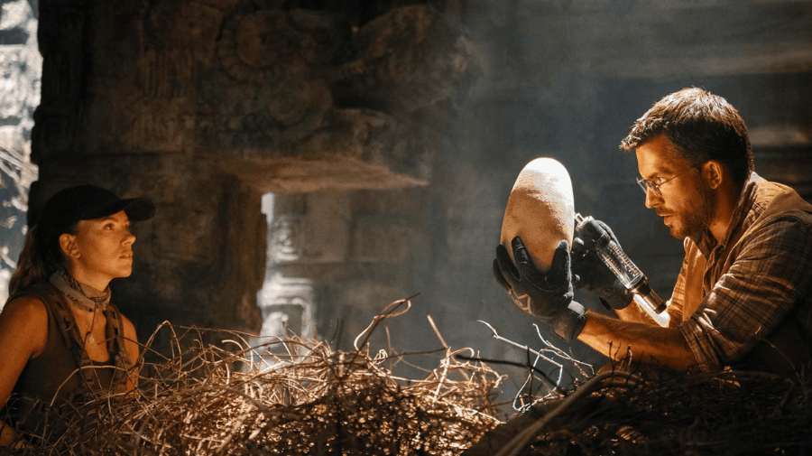 a woman in cap and a man with dark hair and glasses sit either side of a massive bird's nest made of sticks. The man holds a huge egg, in his gloved hands 