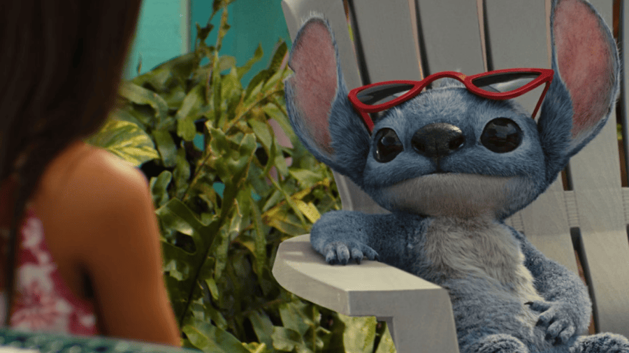 Stitch, a blue alien creature, sits on a sun lounger wearing red sunglasses on his head. Lilo, a young girl with long dark hair, has her back to the camera. 