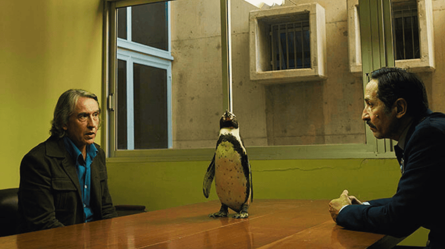 a penguin sits atop a wooden table in a police interview room with yellow walls. One white man with long-ish grey hair sits on the left side of the penguin and a man with short dark hair sit on the right of the table. 