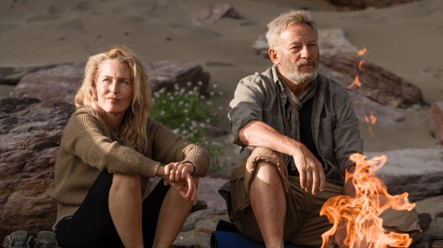 a white man with grey hair and beard and a white woman with long light blonde hair, both dressed in wet weather walking gear and carrying rucksacks, cross grassland.Behind them, cliffs and the sea 