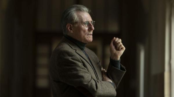 Gabriel Byrne, as Samuel Beckett, looks out of a window. He's wearing a brown-ish tweed suit and glasses 