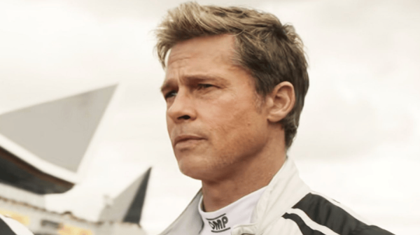 a tanned man with blonde short hair stares off into the distance. We can see he's wearing a racing driver suit, in white, with black stripes on the shoulder 
