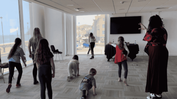 a room with floor to celiling windows down the left side. a group of children and one adult stand with their back to the camera. in the background a few very young children are crawling or sat on the carpet 