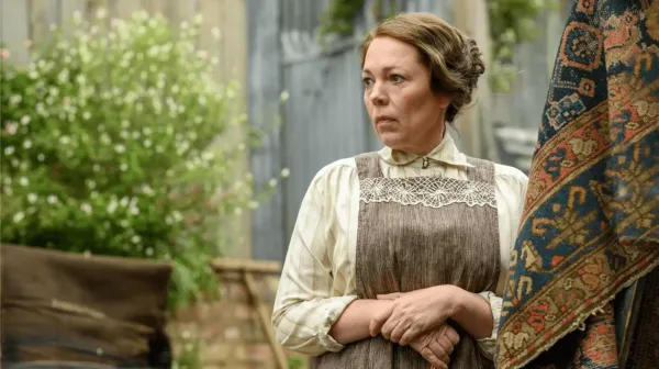Olivia Colman in Edwardian clothes, a pinafore dress and white blouse, stands outside a house. Next to her is a green bush and brickwork