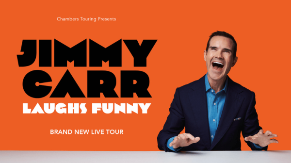 Jimmy Carr wearing a black suit and blue shirt. He's laughing and the background behind is orange.