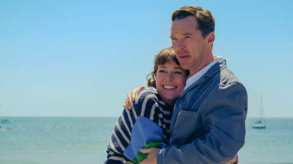 a short, white women with dark bobbed hair and a stripy jumper, is being hugged by a tall white man wearing a light blue jacket. Behind them an idyllic turquoise sea and sky