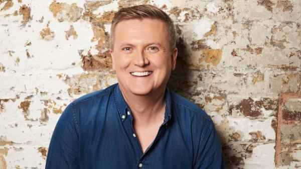 Aled Jones wearing a dark blue shirt, standing in front of a bare-brick wall.