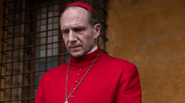Ralph Fiennes, a white male actor in his 60s, is dressed in a Bishop's surplice. It is bright red. Around his neck is a Cruxifix. Behind him, a wall painted ochre yellow 