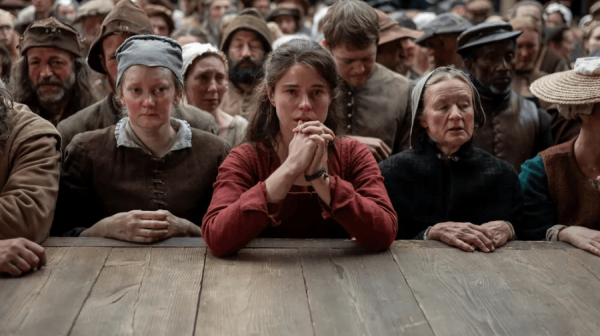 a woman in a 16th century deep red dress, stands wit her elbows on the edge of a stage. she is surrounded by the rest of the audience, all in similar dress