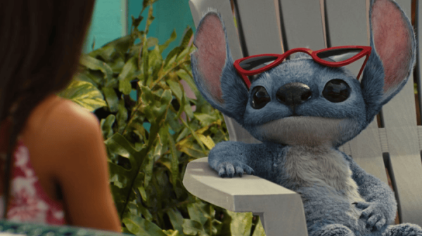 Stitch, a blue alien creature, sits on a sun lounger wearing red sunglasses on his head. Lilo, a young girl with long dark hair, has her back to the camera. 