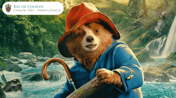 paddington bear is on a raft on a river in peru. He's wearing his duffle coat, waterproof red hat and carrying an umbrella 