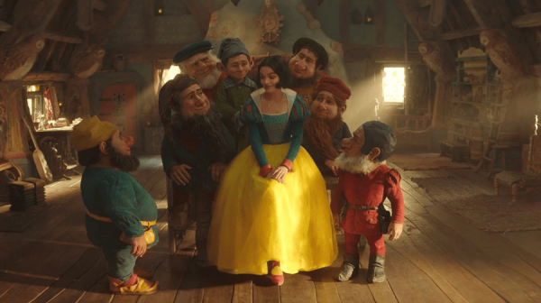 snow white, dressed in a flouncy dress with blue bodice and gold skirt, sits in the cottage of the Seven Dwarfs, surrounded by seven bearded people of small stature with beards, all wearing brightly coloured trousers and tops 