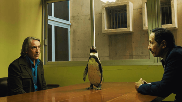 a penguin sits atop a wooden table in a police interview room with yellow walls. One white man with long-ish grey hair sits on the left side of the penguin and a man with short dark hair sit on the right of the table. 