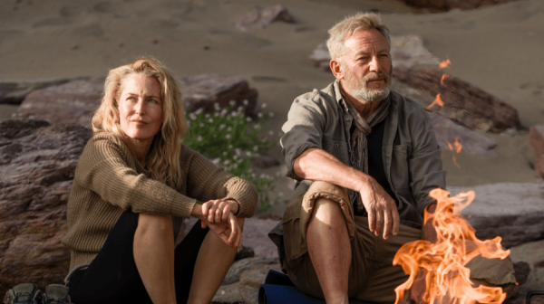 a white man with grey hair and beard and a white woman with long light blonde hair, both dressed in wet weather walking gear and carrying rucksacks, cross grassland.Behind them, cliffs and the sea 
