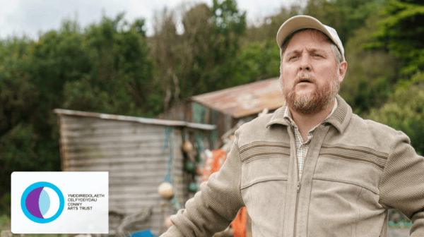 a white man in his 30s with a beard, khaki coat and basecall cap, stands with one hand on a fence looking off camera. He's outside, with a shed and green bushes behind him. There's a logo for Conwy Arts Trust in the bottom left of the image